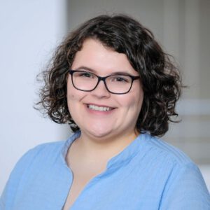 Lächelnde Person mit kurzen lockigen Haaren und Brille in einem hellblauen Hemd im Innenbereich.