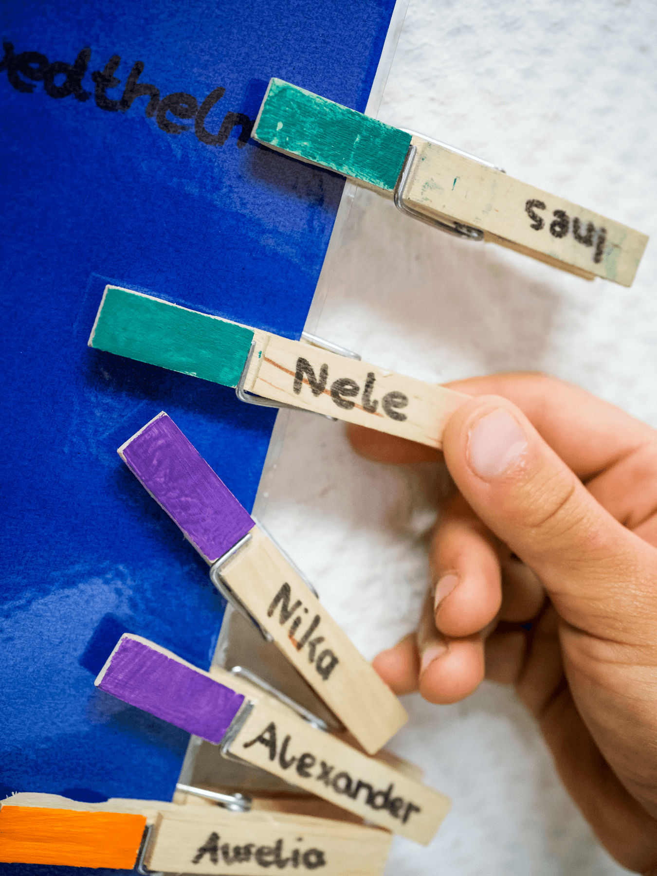 Eine Hand schnappt sich eine Wäscheklammer mit der Aufschrift „Nele“ aus einer Reihe beschrifteter Holzwäscheklammern mit unterschiedlichen Farben an der Spitze, die an einem blauen Brett befestigt sind. Auf anderen Klammern stehen die Namen „Savi“, „Nika“, „Alexander“ und „Aurelia“.