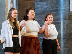 Drei junge Frauen stehen nebeneinander in einem Gebäude, halten sich an den Händen und blicken nach vorn. In ihrer legeren Kleidung wirken sie vor dem Hintergrund grauer Steinmauern vereint und selbstbewusst.