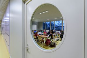 Blick durch ein Bullauge in das Unterrichtsgeschehen eines Klassenraumes.
