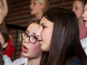 Zwei Mädchen singen gemeinsam im Chor. Eines trägt eine Brille. Im unscharfen Hintergrund sind weitere Personen zu sehen. Hinter ihnen befindet sich eine Backsteinmauer.