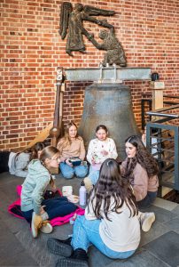 Eine Gruppe von sechs Mädchen sitzt im Kreis auf dem Boden, unterhält sich und isst vor einer großen Glocke, die unter einer Wandskulptur eines Engels und einer knienden Figur vor einer roten Backsteinwand angebracht ist.