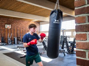 Ein junger Mann mit roten Boxhandschuhen und Sportkleidung schlägt in einer geräumigen Turnhalle auf einen hängenden Boxsack ein. Im Hintergrund sind Fitnessgeräte und Ziegelwände zu sehen.