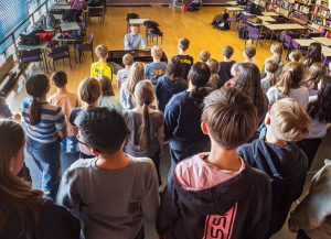 Eine große Gruppe von Schülern steht in einer geräumigen Bibliothek oder einem Klassenzimmer mit Bücherregalen und Tischen vor einem Klavier spielenden Mann. Die Schüler sind von hinten zu sehen.