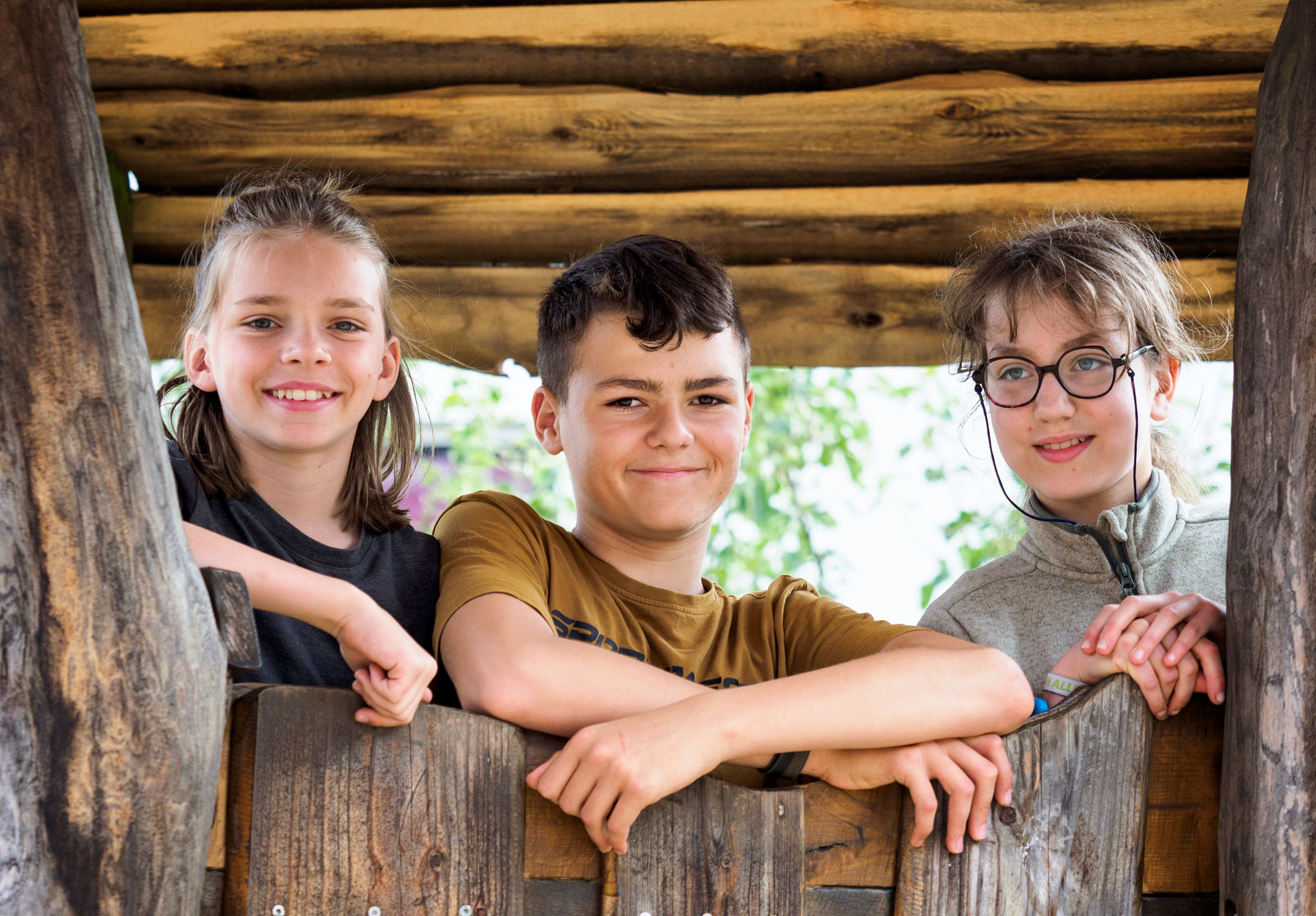 Drei lächelnde Kinder lehnen an einem Holzzaun im Freien, unter einem begrünten Unterstand.