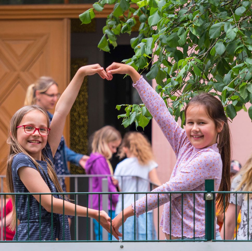 Zwei lächelnde Mädchen bilden draußen ein Herz mit ihren Armen; andere Kinder und ein Erwachsener stehen hinter ihnen.
