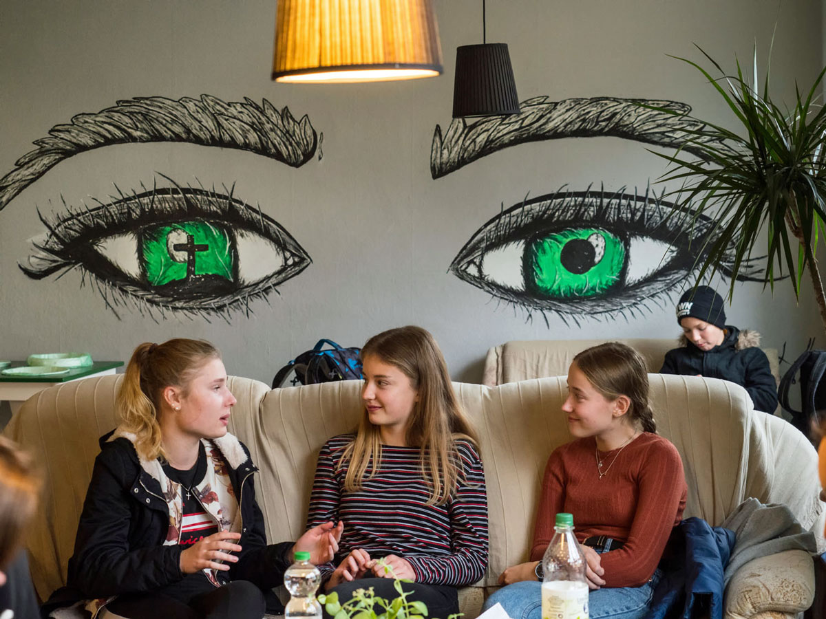 Drei Mädchen unterhalten sich auf einer Couch; ein Junge im Hintergrund. Eine Wandmalerei mit grünen Augen ziert die Wand.