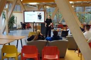 Eine Frau hält einen Vortrag vor Schülern in einem modernen Klassenzimmer; die Folie zeigt "Wie die Gesellschaft die Frauen sah" und ein historisches Foto.