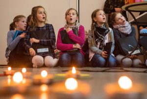 Fünf junge Mädchen sitzen im Schneidersitz und singen mit Mappen, vor ihnen leuchtende Kerzen.