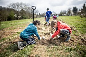 Zwei Kinder pflanzen in einem Garten mit mit Schnüren markierten Reihen; andere arbeiten in der Nähe unter bewölktem Himmel.
