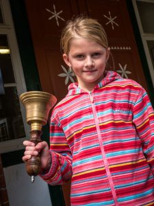 Mädchen in gestreifter Jacke hält eine Messinghandglocke vor einer braunen Tür mit weißen Sternen.