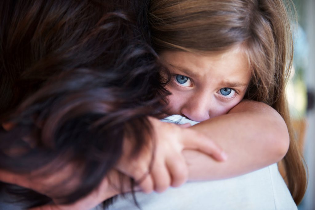 Ein besorgtes junges Mädchen mit blauen Augen umarmt einen Erwachsenen, dessen Gesicht nicht zu erkennen ist.