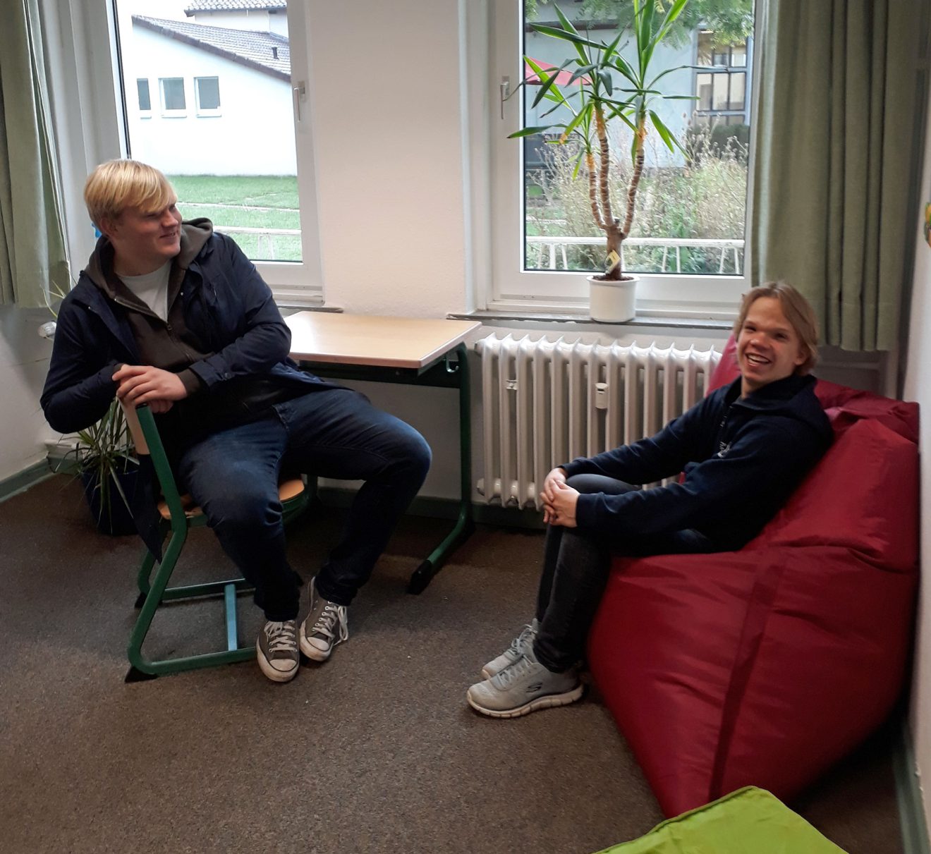 Zwei Menschen unterhalten sich und lächeln in einem Raum mit einem grünen Stuhl, einem roten Sitzsack und einem Fenster. Bild: Paul-Gerhardt-Schule