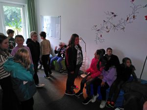 Kinder in einem Klassenzimmer mit einer blauen Bank, einem Whiteboard, einer Vogelwand und Tageslicht durch grüne Vorhänge. Bild: Paul-Gerhardt-Schule