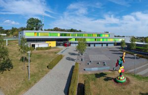 Modernes Gebäude mit grünen und gelben Tafeln, Bäumen, einer bunten Skulptur und Gehwegen.