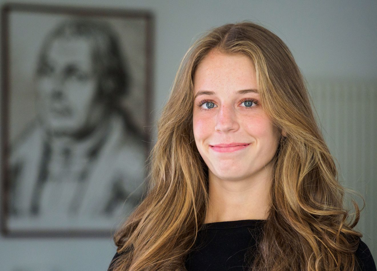 Junge blonde Frau in schwarzem Oberteil lächelt in die Kamera; hinter ihr ist ein verschwommenes Porträt zu sehen.