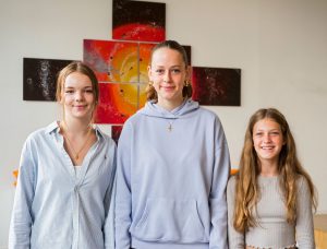 Drei lächelnde Mädchen stehen drinnen vor einem kreuzförmigen bunten abstrakten Gemälde. Foto: Martin Kirchner.