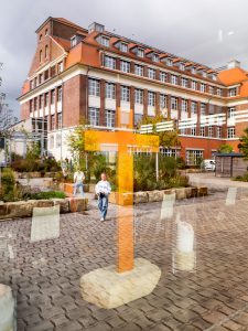 Rotes Backsteingebäude mit vielen Fenstern, orangefarbener Kunst und zwei Personen auf Bänken im Freien.