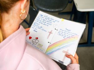 Person im rosa Kapuzenpulli zeigt ein Notizbuch mit deutschen Gebeten und bunten, kindlichen Zeichnungen.