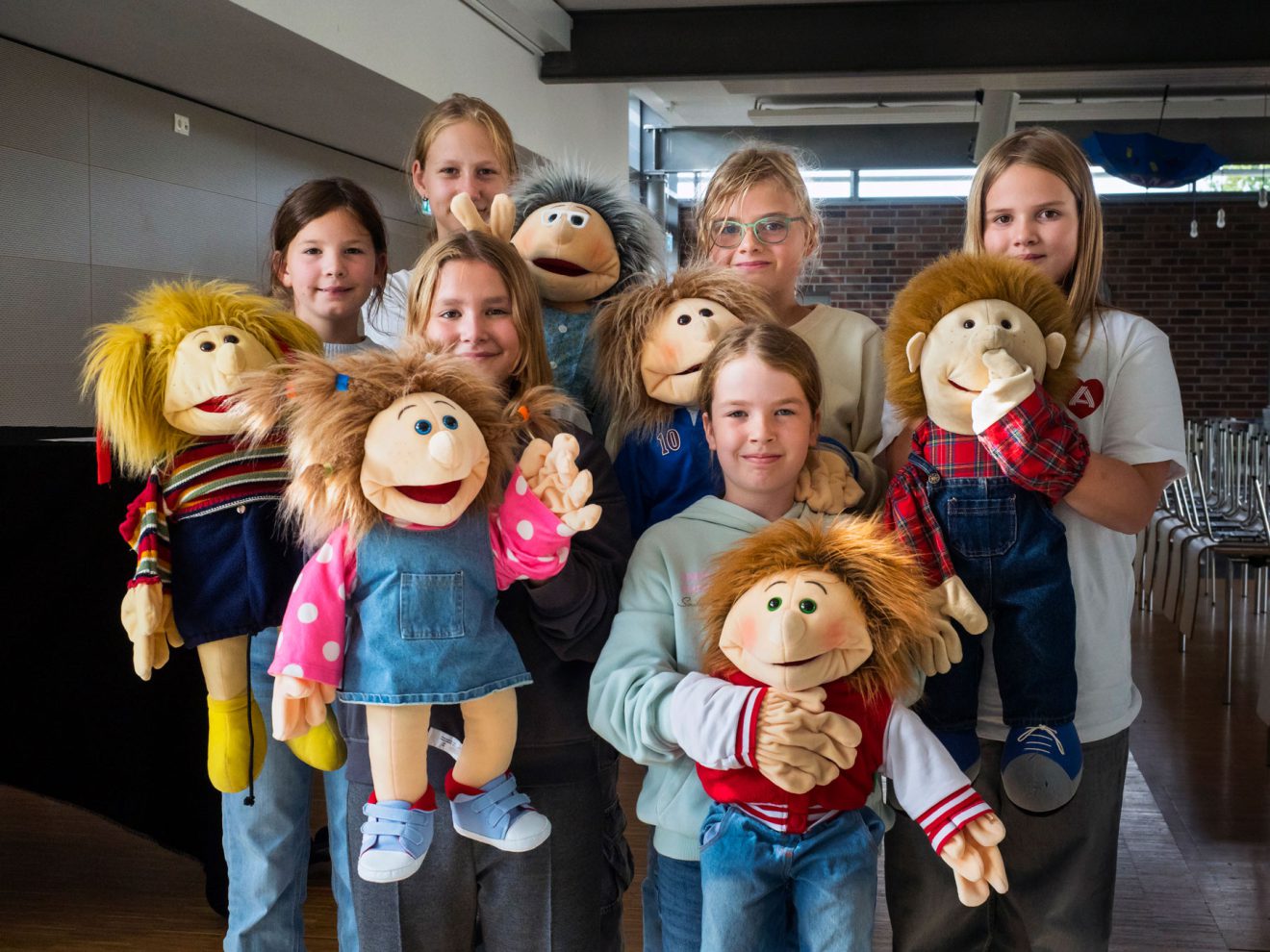 Sieben lächelnde Kinder halten bunte Handpuppen und stehen in einem hellen Innenraum zusammen.