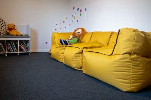 Ein gemütliches Zimmer mit gelben Sitzsäcken, einer Puppe, einem kleinen Bücherregal und bunten Wandtupfen.