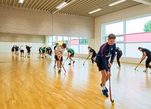 Junge Leute üben in einer Turnhalle Feldhockey, jeder mit einem Schläger und einem Ball.