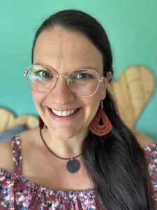 Frau mit langen dunklen Haaren und Brille lächelt, trägt ein geblümtes Oberteil und orangefarbene Ohrringe.