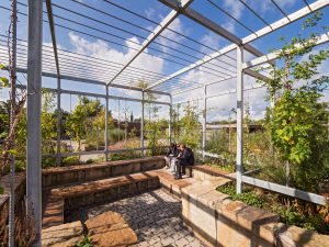 Zwei Personen sitzen auf einer Holzbank unter einer Metallpergola, umgeben von Pflanzen und Steinmauern.