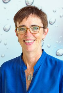 Frau mit kurzen braunen Haaren und Brille, in einer blauen Bluse, lächelt vor einem blauen Hintergrund.