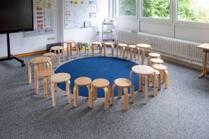 Ein Klassenzimmer mit kreisförmig angeordneten Holzhockern auf einem blauen Teppich, erhellt von natürlichem Licht.