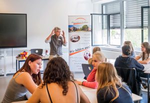 Der Lehrer spricht zu den Schülern in kleinen Gruppen; dahinter Banner, Leinwand und offene Fenster.