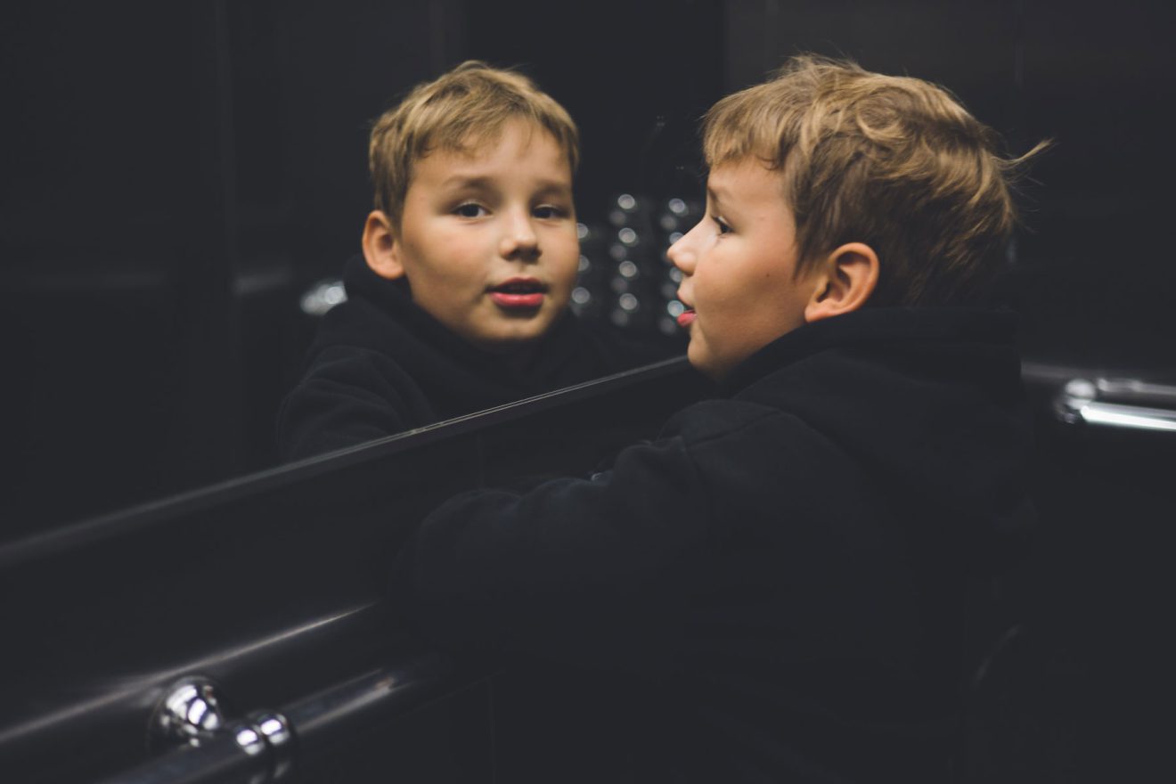 Ein Junge in einem schwarzen Kapuzenpulli steht vor einem Spiegel; sowohl sein Gesicht als auch sein Spiegelbild sind bei schwachem Licht sichtbar.