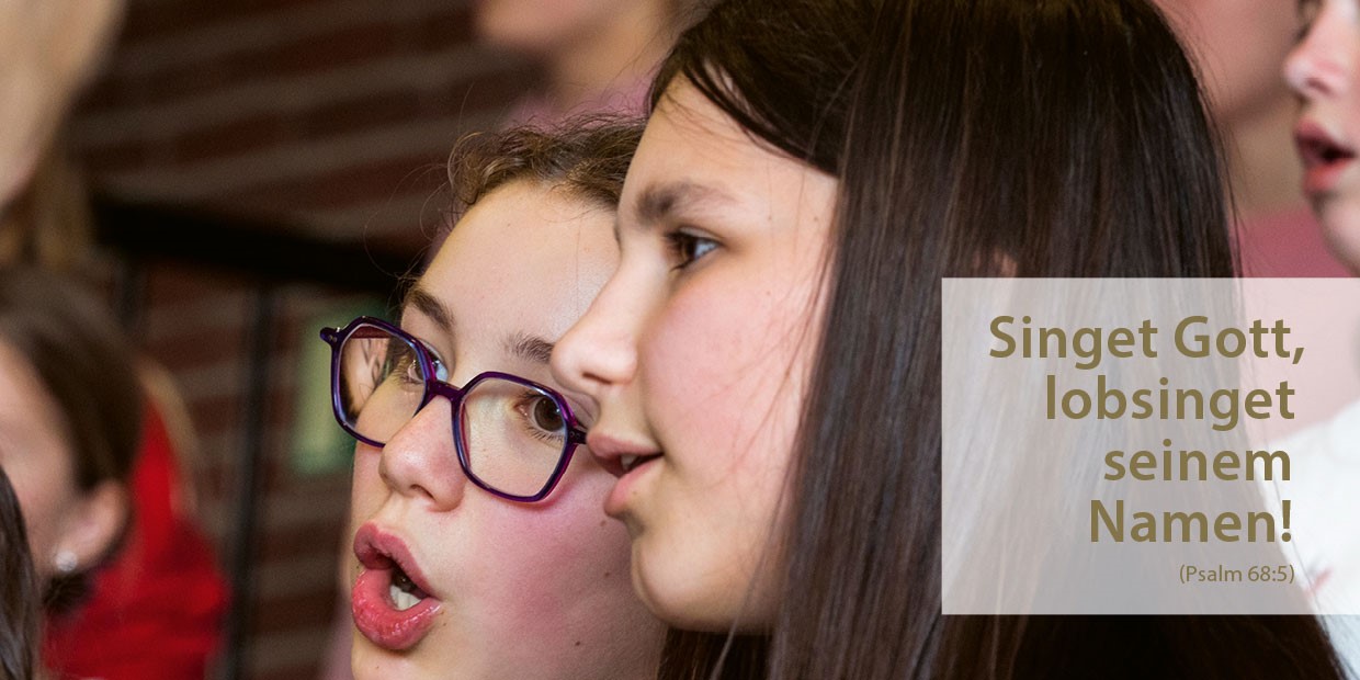 Zwei fröhliche Mädchen singen im Chor; der deutsche Text neben ihnen zitiert Psalm 68,5.