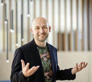 Lächelnder Mann mit Glatze und Bart gestikuliert in Blazer und geblümtem Hemd vor Holzpaneelen.