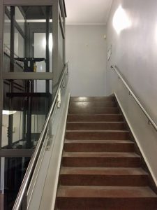 Eine braune Treppe mit Metallschienen verläuft neben einem gläsernen Aufzug in einem Gebäude mit grauen Wänden.