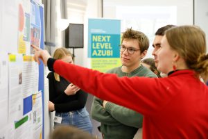 Frau in rotem Pullover überreicht einer Gruppe Geschenke; im Hintergrund das Schild "NEXT LEVEL AZUBI".