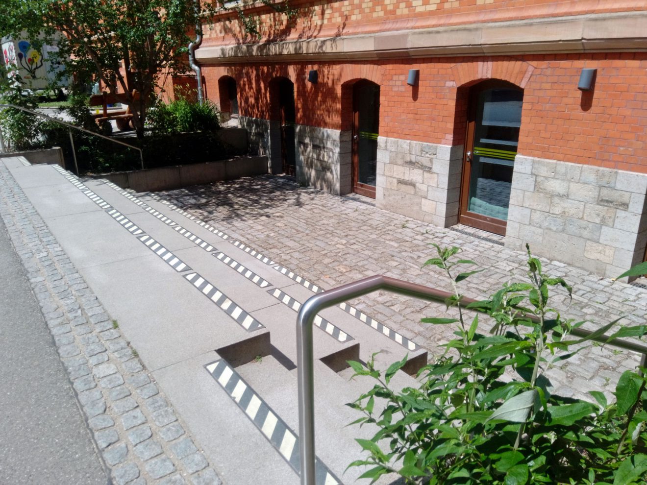 Treppen und Rampen an einem roten Backsteingebäude, mit gestreiften Kanten, Pflanzen und Handläufen.
