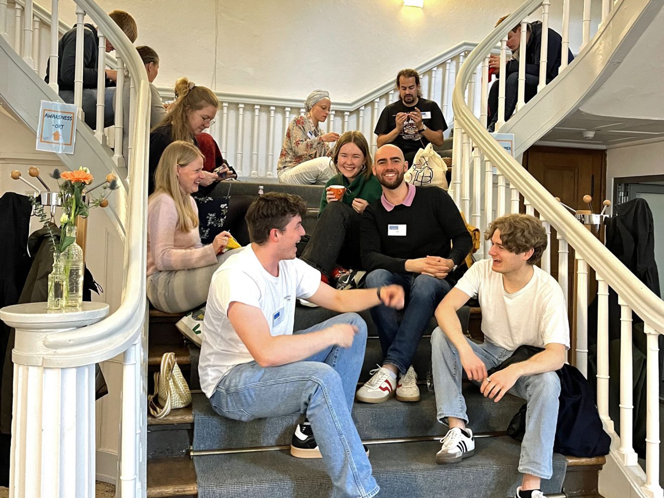 Die Menschen sitzen auf einer Treppe in einem Innenraum, lächeln und unterhalten sich mit Getränken in der Nähe von Blumenvasen. Bild: Evangelisches Studienwerk