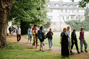 Menschen gehen auf einem von Bäumen gesäumten Parkweg in der Nähe eines großen Gebäudes mit vielen Fenstern.