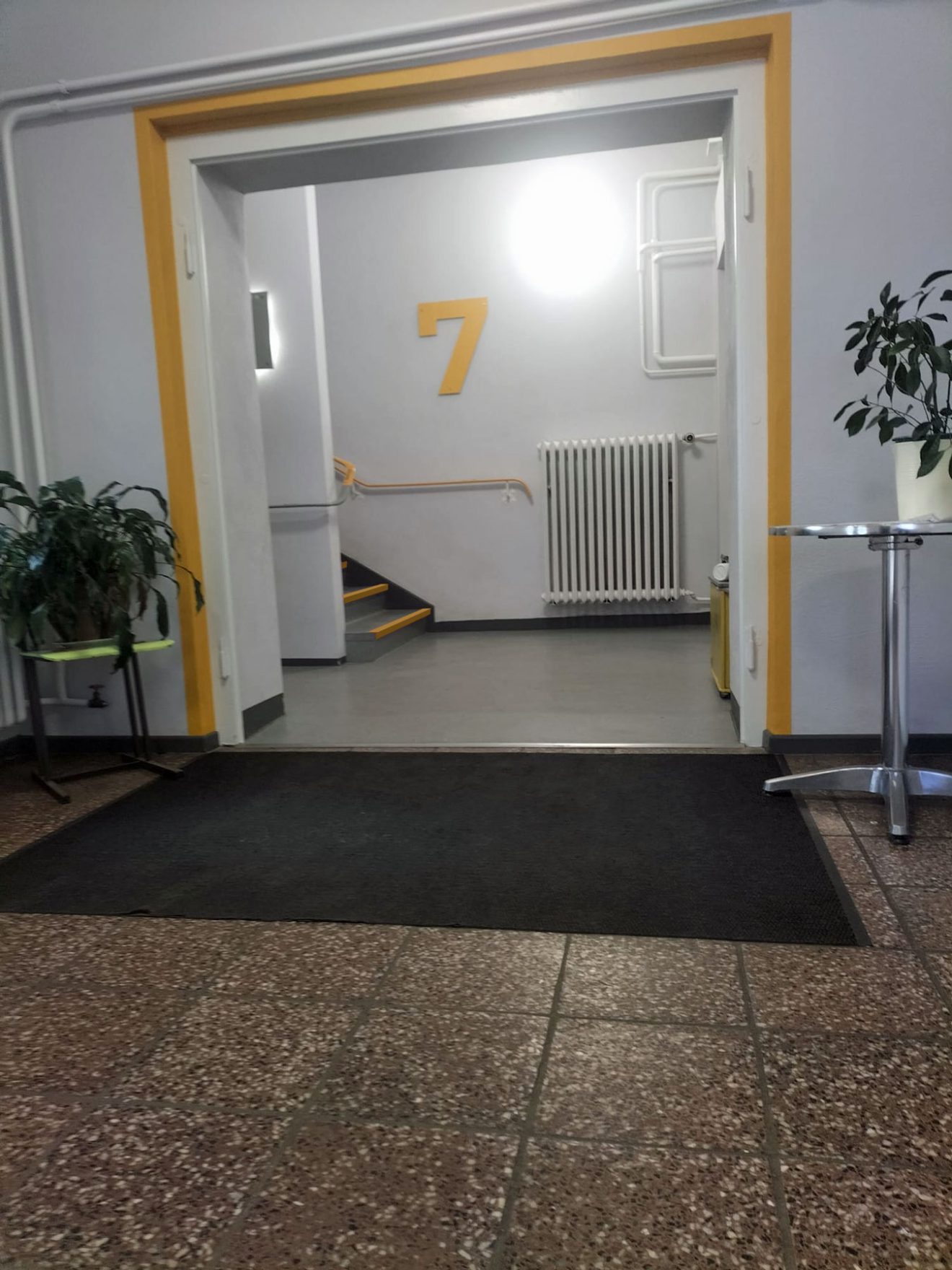 Flur mit einer gelben Nummer 7 über einem Heizkörper, Treppe, Pflanzen und einem kleinen Tisch.