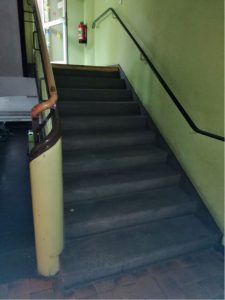 Schummriges Treppenhaus mit schwarzen Stufen, hellbraunem Handlauf, Feuerlöscher und Licht von der Tür aus.