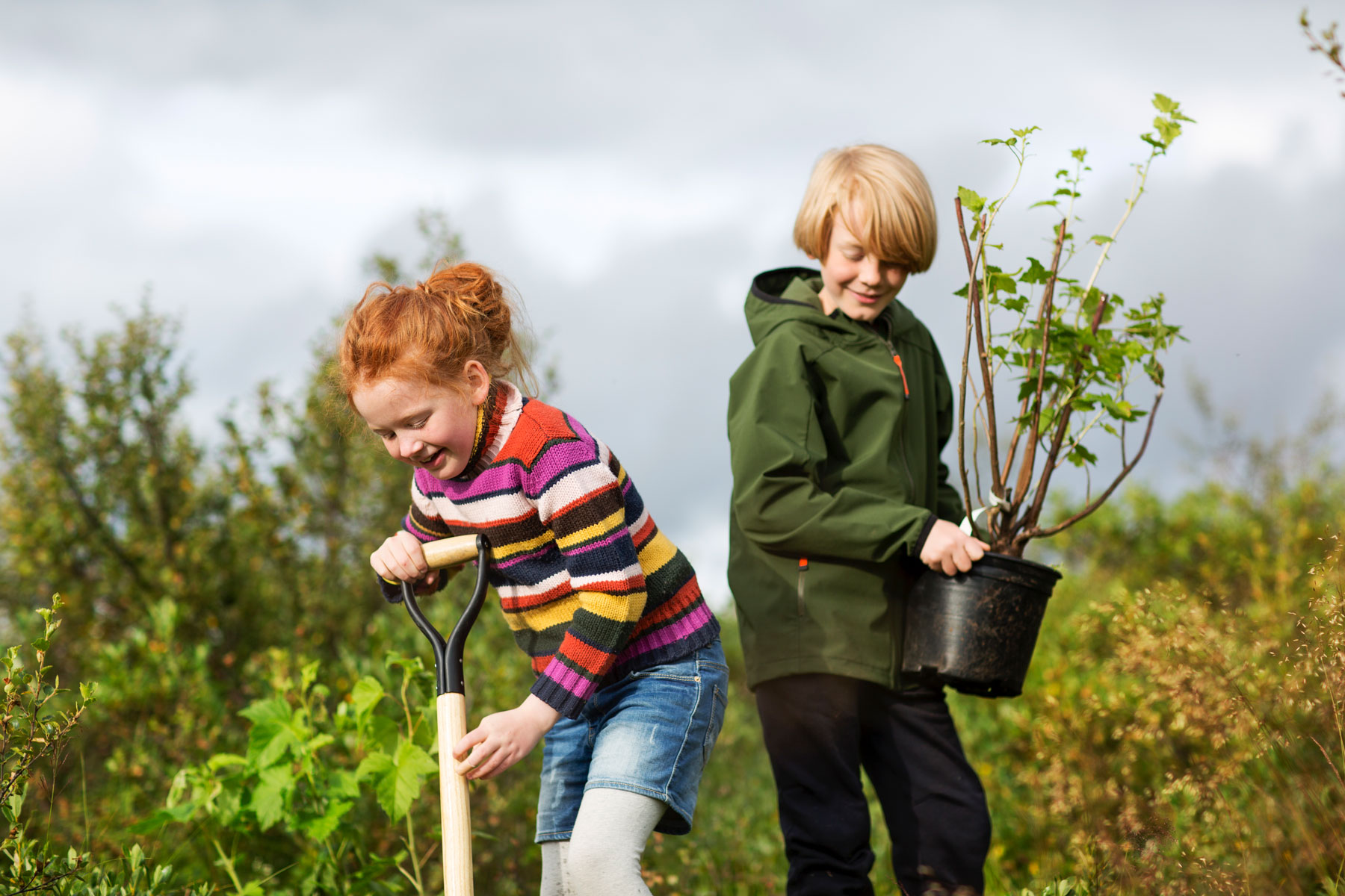 Zwei Kinder im Freien: ein rothaariges Mädchen gräbt, während ein blonder Junge eine Topfpflanze hält.