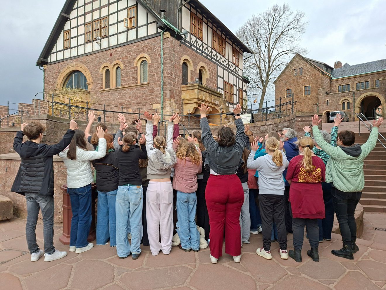 Vor einem historischen Gebäude aus Stein und Holz heben die Menschen in einem Halbkreis die Hände.