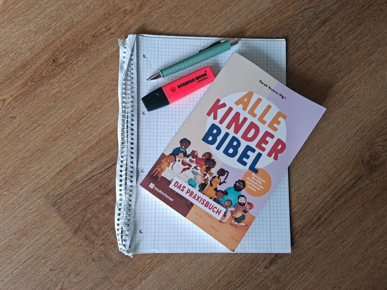 Notizbuch, grüner Stift, rosa Textmarker und "Alle Kinder Bibel Praxisbuch" auf Holztisch.