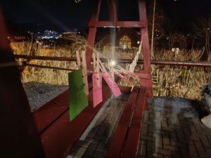 Rote Holzschaukel bei Nacht mit bunten Karten, die an einer Schnur befestigt sind; dahinter Stadtbeleuchtung und Haus.