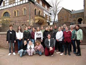 Jugendliche und Erwachsene posieren auf den Steinstufen vor einem historischen Backsteingebäude mit Rundbogenfenstern.