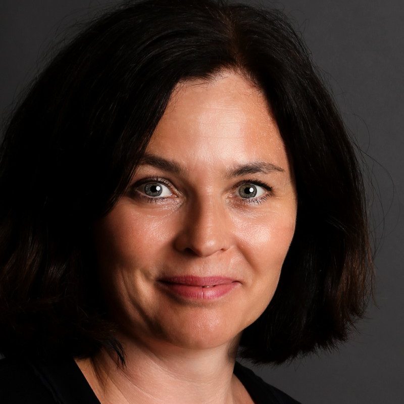 Frau mit dunkelbraunem Haar und hellen Augen posiert in einem schwarzen Oberteil vor einem dunklen Hintergrund.