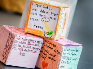 Drei bunte Papierwürfel mit deutschen Gebeten und Zeichnungen sind auf einem Tisch gestapelt. Bild: Martin Kirchner.