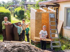 Ein Mann und vier Kinder bauen in einem sonnigen Garten einen kleinen Holzschuppen und halten dabei Bretter und Planken fest. Foto: Martin Kirchner