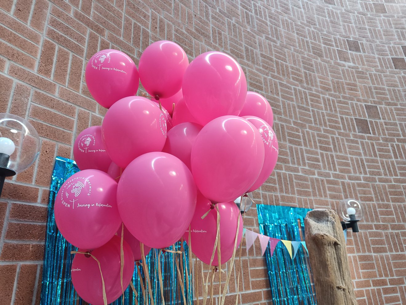 Rosa Luftballons mit aufgedruckten Motiven schweben im Innenraum in der Nähe einer Backsteinwand und einer Lichterkette. Foto: Janne Neumann